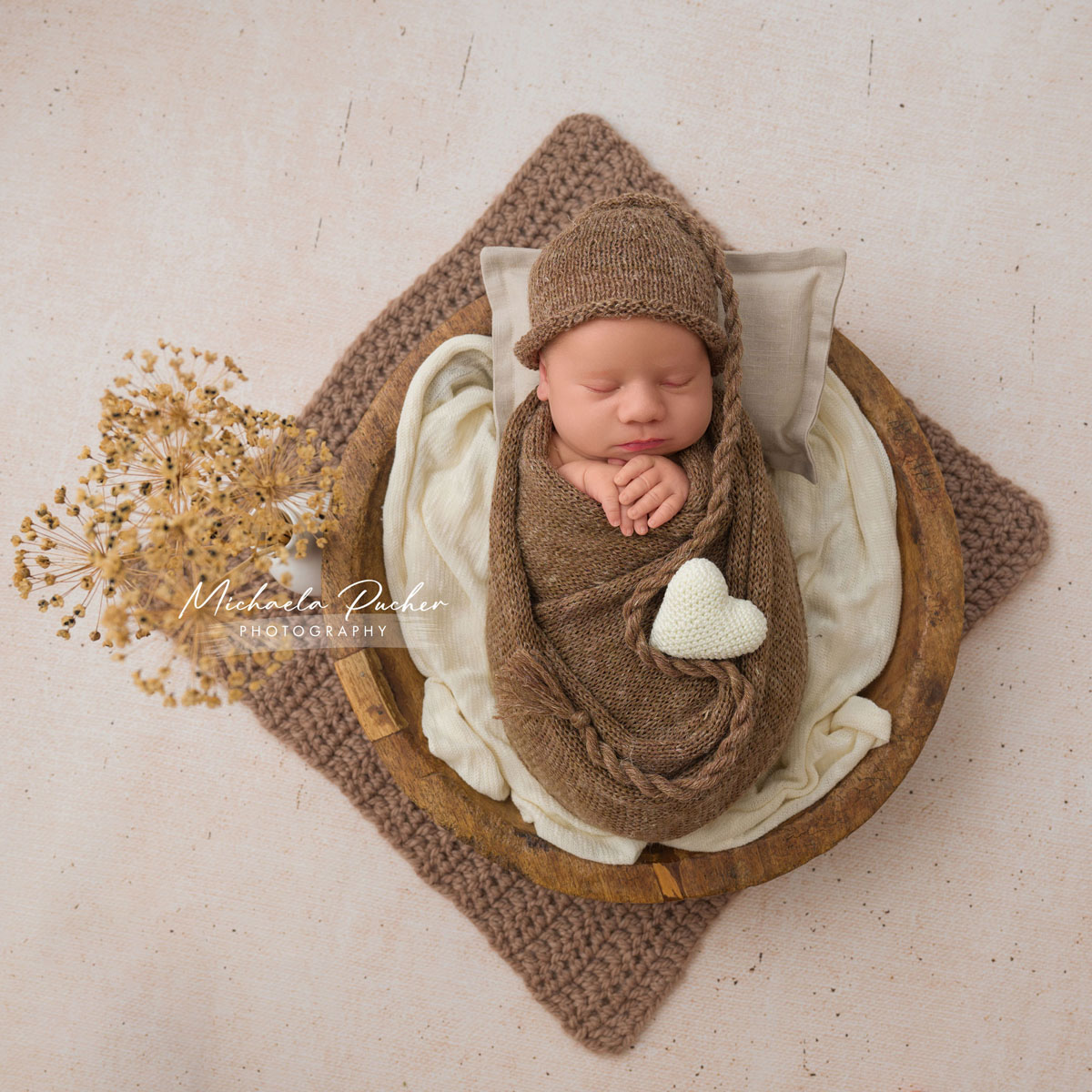 Neugeborenes schläft eingewickelt in braunen Strickstoffen in einer runden Holzschale, dekoriert mit einem weißen Häkelherz und getrockneten Blüten, auf einer beigefarbenen Unterlage.